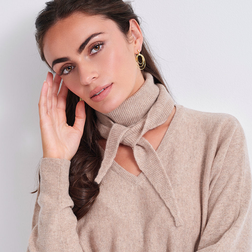 A woman wearing a beige knit sweater with a high ribbed turtleneck collar and a front tie detail, styled with gold hoop earrings against a light gray background.
