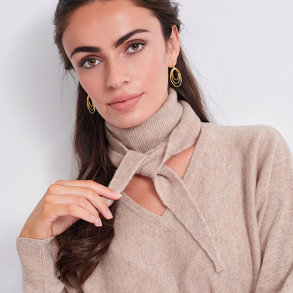 A close-up of a woman wearing a beige knit turtleneck sweater with a tie-front cutout and gold hoop earrings, photographed against a plain light background.