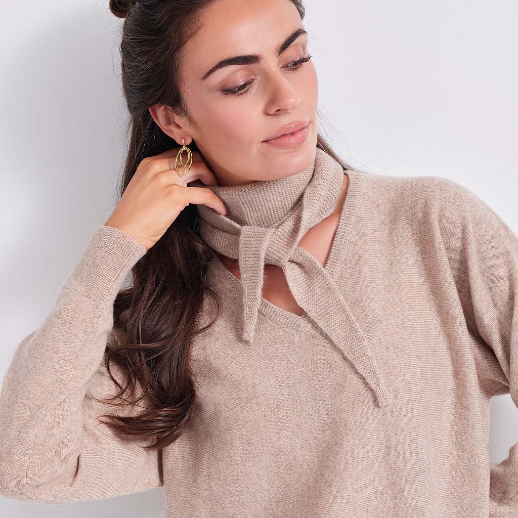 A woman wearing a beige knit sweater with a tie-neck detail and a matching beige knit neck scarf, paired with gold hoop earrings against a plain light background.