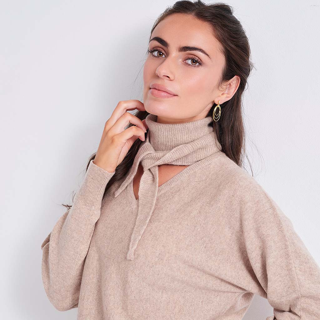 A woman wearing a beige knit sweater with a high folded collar and a front keyhole cutout tied with a matching cord, posed against a plain light gray background.