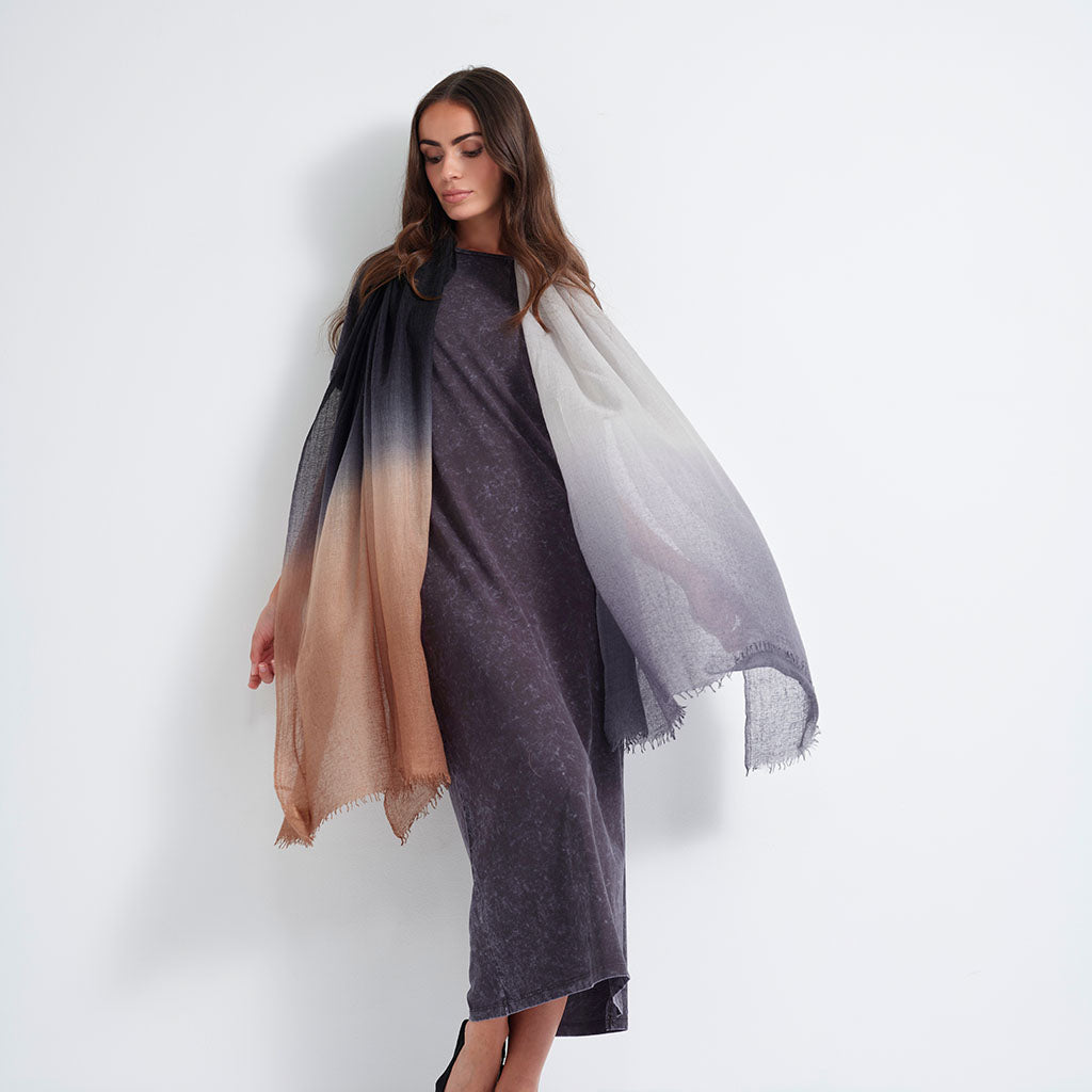 A woman models a lightweight ombre shawl draped over her shoulders. The shawl features a gradient transition from black at the top, through taupe-brown in the middle, to off-white at the bottom.