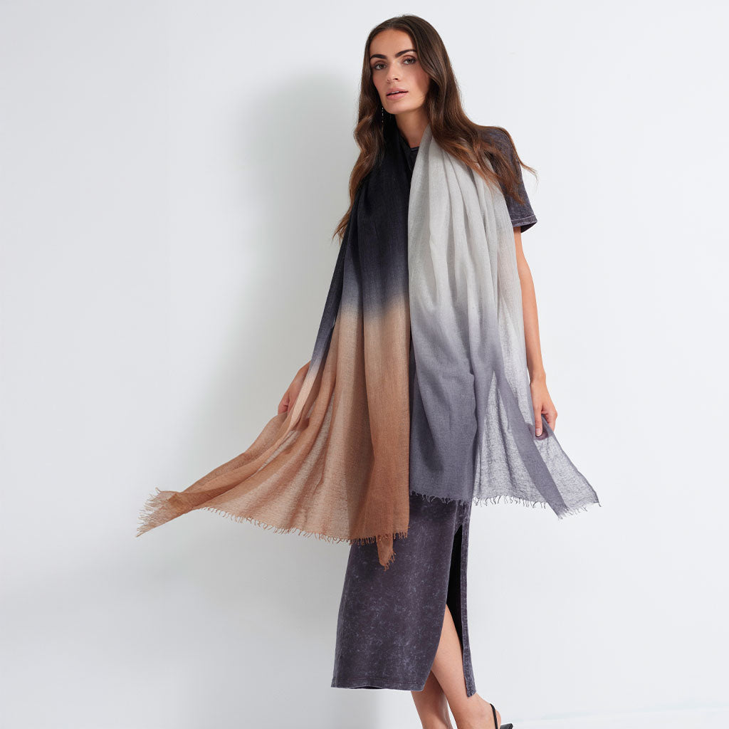 A woman models a lightweight ombre scarf with fringed edges, featuring a color gradient from black, through gray, to a warm tan-brown. She is wearing a black t-shirt and a gray midi dress against a plain white studio background.
