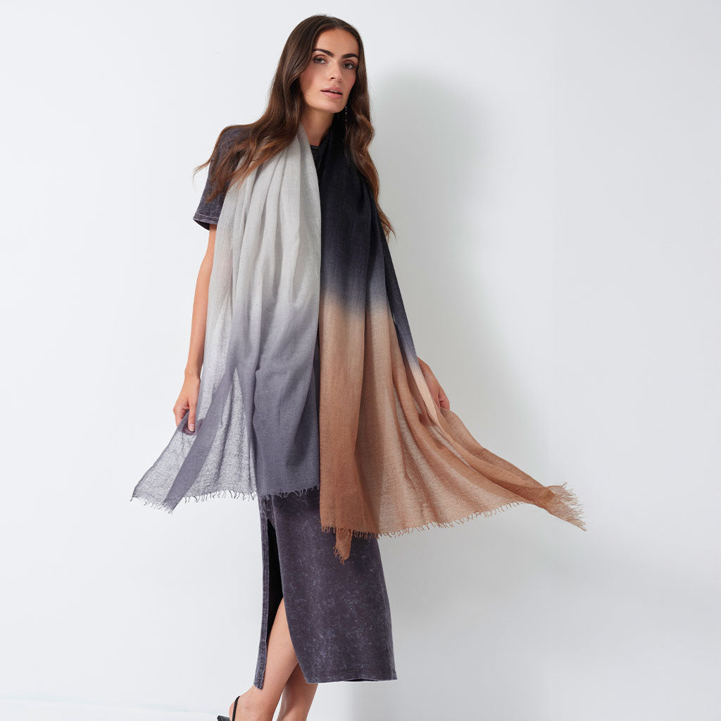 A woman models a lightweight gradient shawl draped over her shoulders, transitioning from light gray at the top through black in the middle to a warm tan/beige at the bottom, paired with a dark gray midi dress against a plain white studio background.