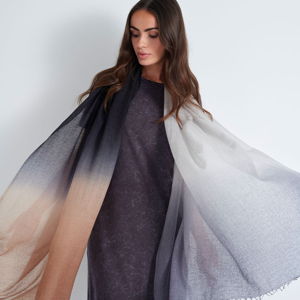 A woman wearing a dark purple textured dress, draped in a large semi-transparent ombre shawl that transitions from black to beige and then to light gray, against a plain light gray background.