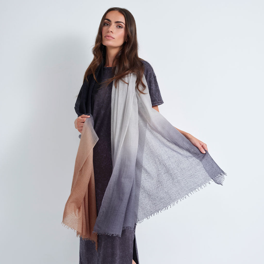 A woman modeling a lightweight, semi-sheer shawl with a gradient color story, featuring a dusty blue-gray to light gray ombré pattern, styled over a dark gray tunic dress. She holds one side of the shawl to show its texture and fringe detail.