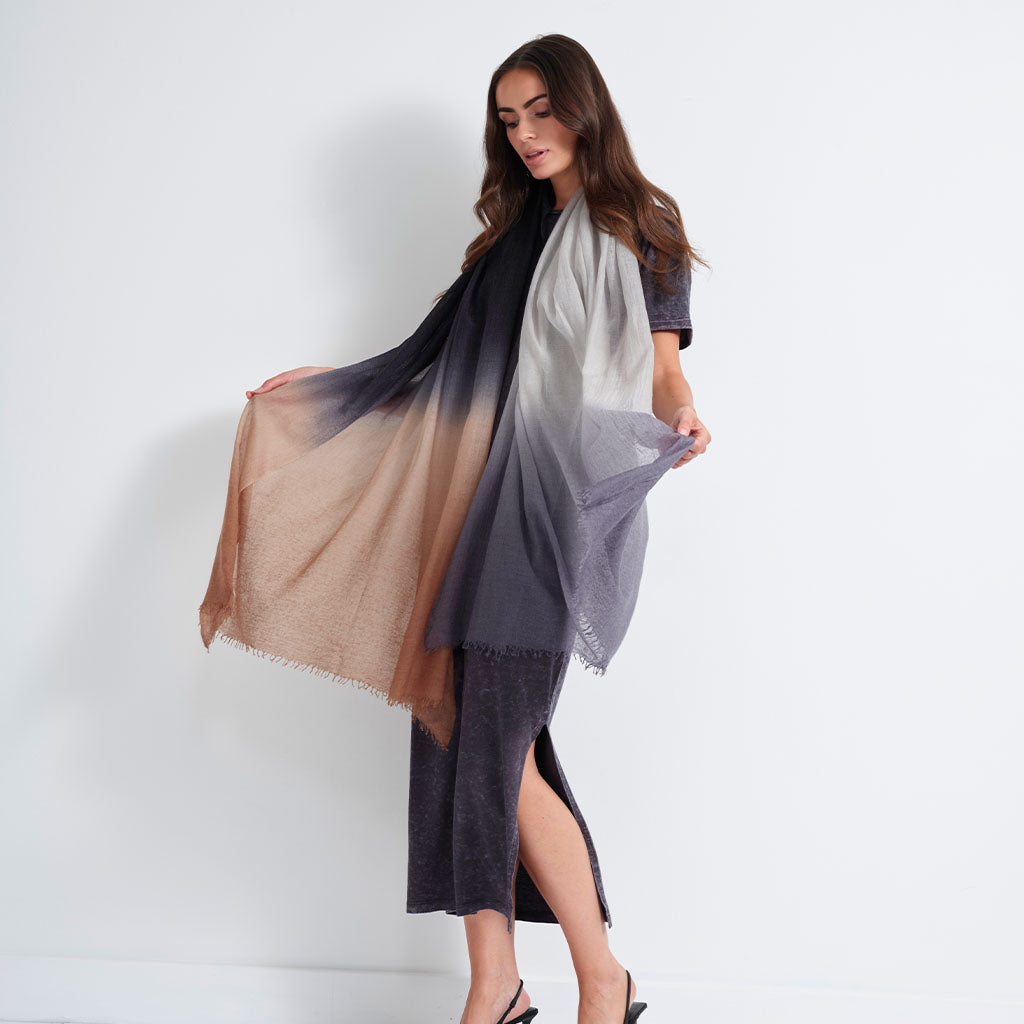 A woman models a lightweight gradient shawl draped over her shoulders, transitioning from a warm tan/beige on one side through white in the center to a dark charcoal gray on the other. She is wearing a dark gray midi dress and black heels, standing against a clean white studio background.