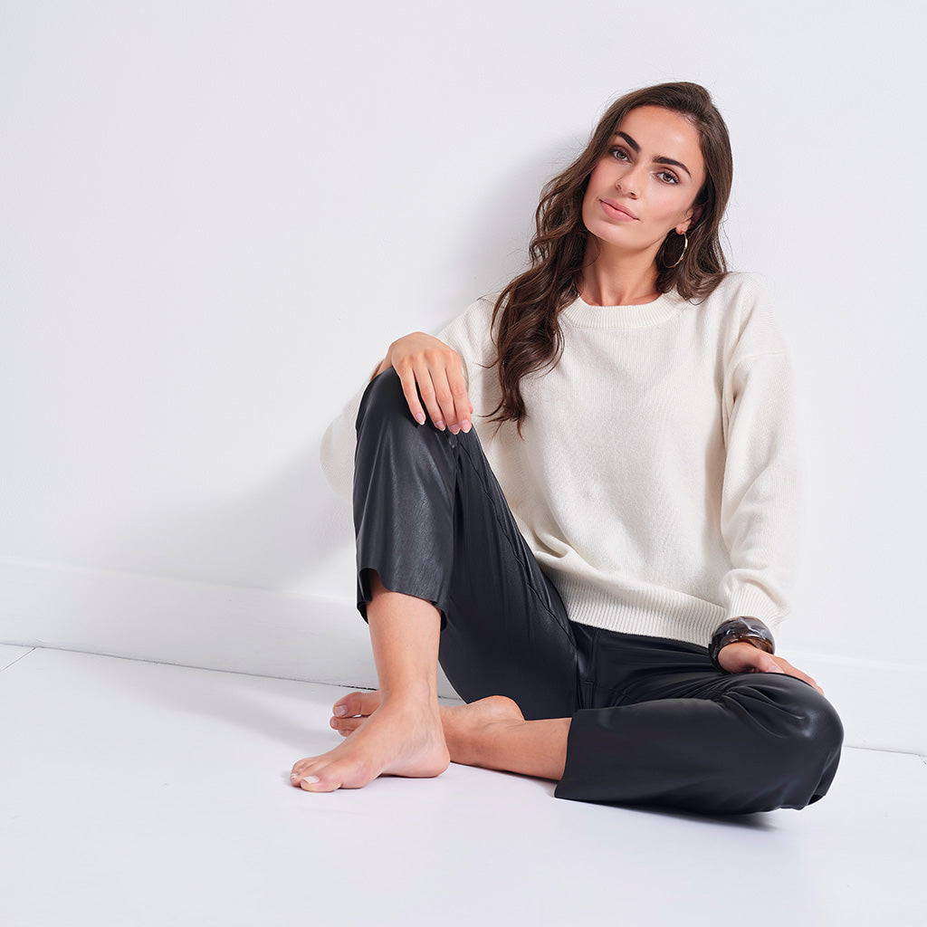 A woman sits cross-legged on a white floor against a white wall, wearing a cream knit crewneck sweater and black leather cropped pants.