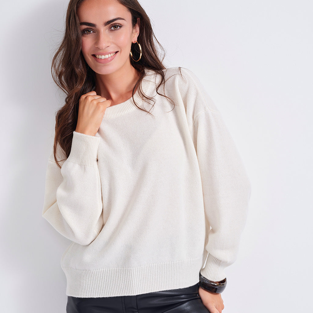 A woman wearing a cream-colored knit crewneck sweater, paired with black leather pants, against a plain white background.