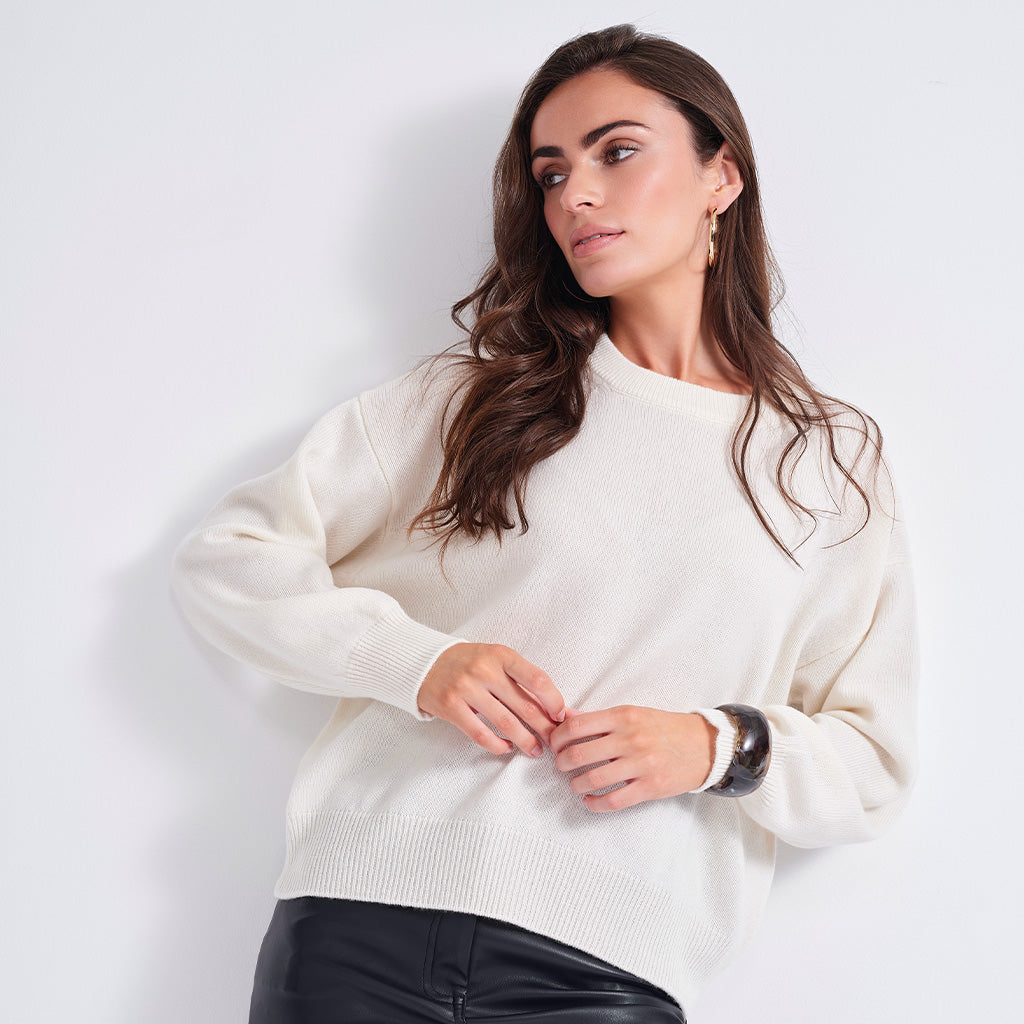 A woman wearing a cream-colored knit crewneck sweater and black pants, posing against a light gray studio background.