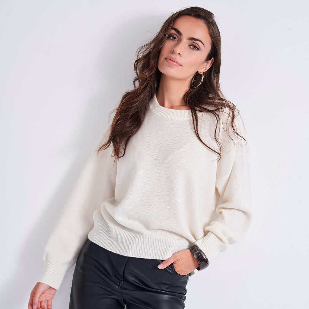 A woman wearing a cream-colored knit crewneck sweater and black leather pants, posing against a light gray studio background.