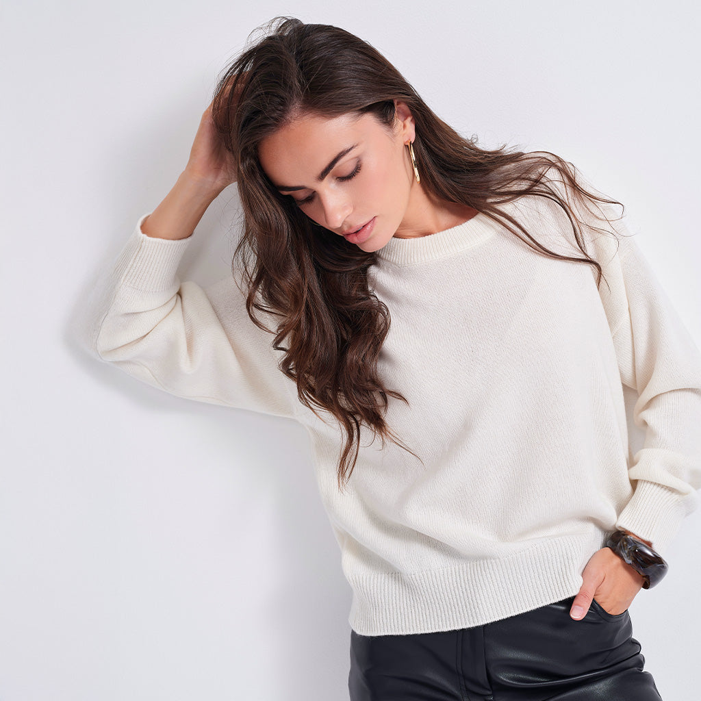 A woman with long wavy brown hair wearing a cream-colored knit crewneck sweater and black leather pants, posing against a light gray studio background.