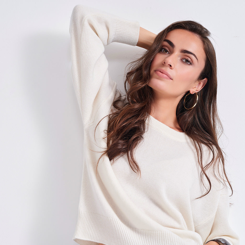 A woman with long wavy brown hair wearing a cream-colored knit sweater and large gold hoop earrings, posing against a plain white wall.