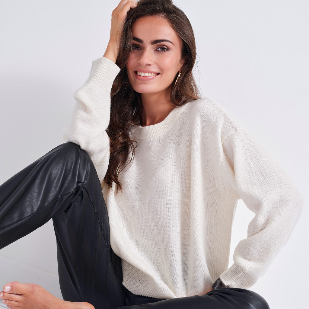 A woman wearing a cream-colored knit crewneck sweater and black pants, sitting against a light gray background.
