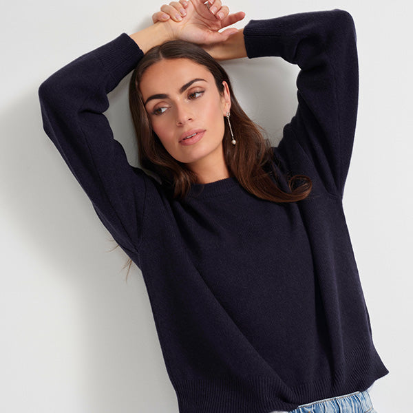 A woman wearing a dark navy blue knit sweater, posing with her arms raised against a plain light gray background.