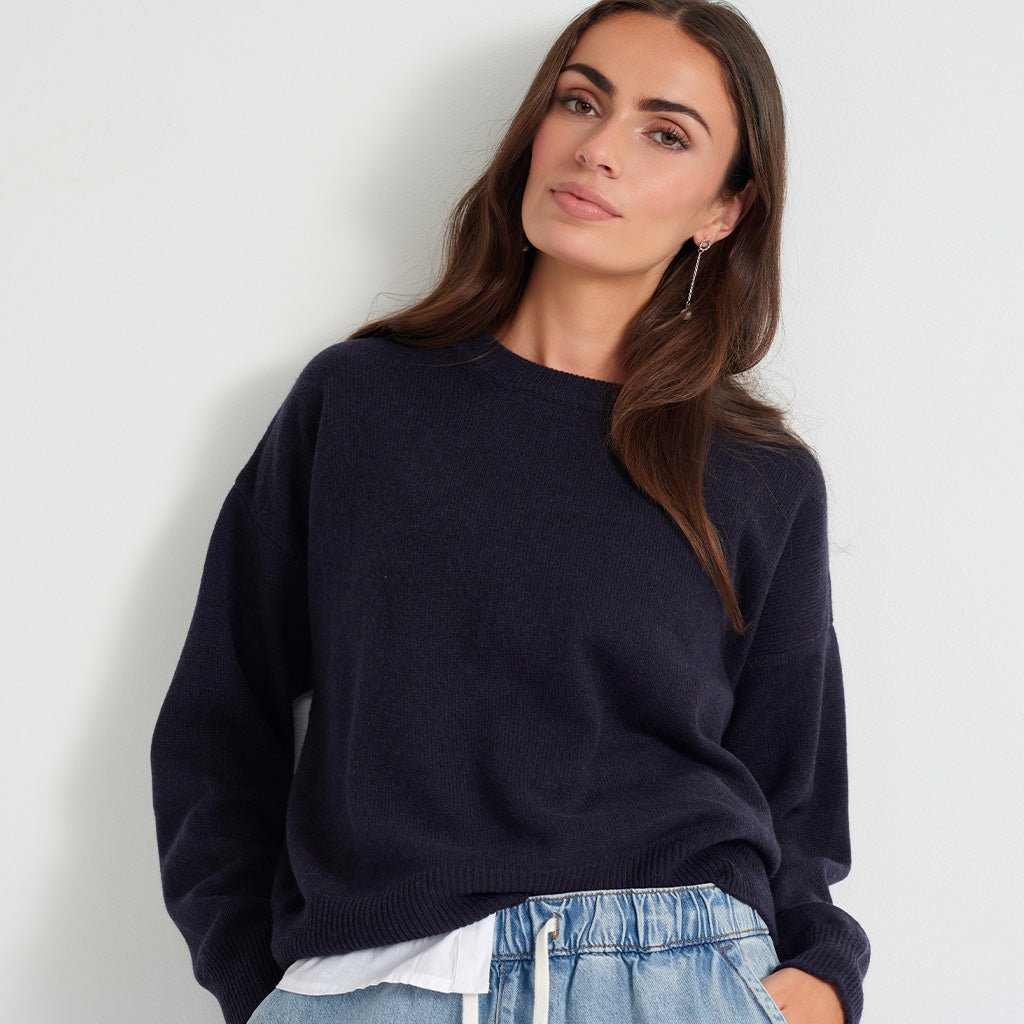 A woman wearing a dark navy blue crewneck knit sweater and light blue denim shorts, posing against a plain light gray wall.