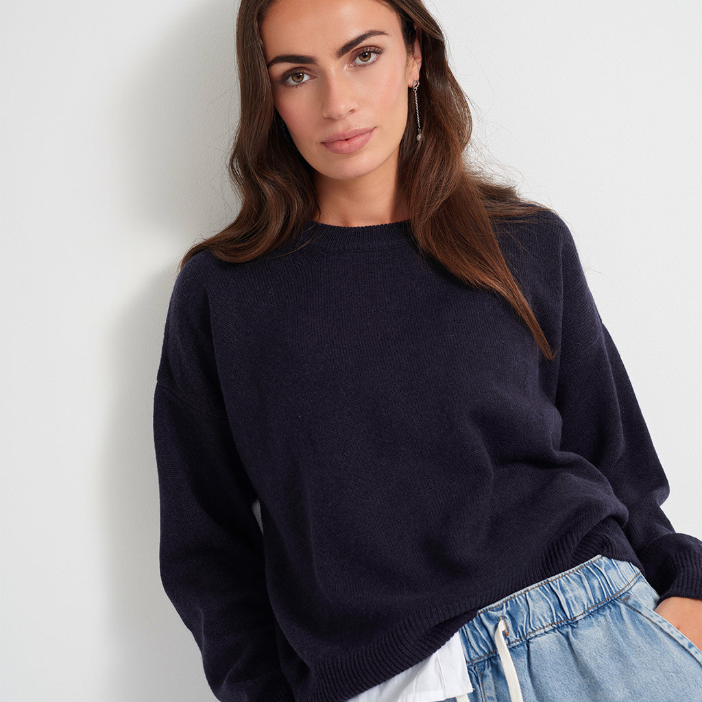 A woman wearing a dark navy blue knit crewneck sweater and light blue denim shorts, posing against a plain off-white wall.