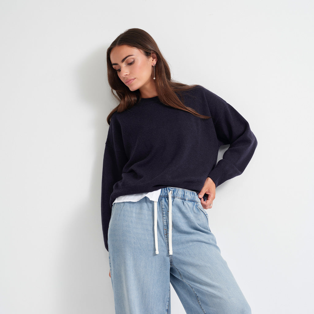 A woman wearing a dark navy blue knit sweater and light blue wide-leg jeans with a white drawstring, posing against a plain white wall.