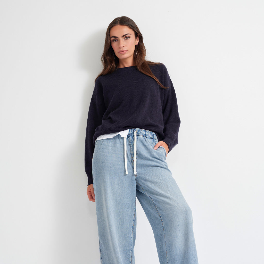 A woman modeling a dark navy crewneck knit sweater and light blue wide-leg jeans with a white drawstring, standing against a plain white wall.