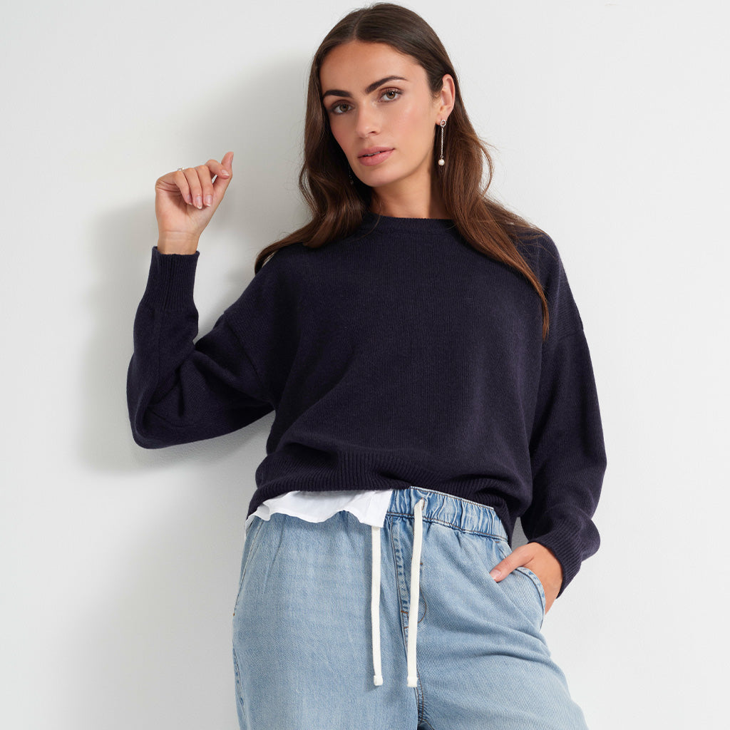 A woman wearing a dark navy blue knit sweater and light blue jeans with a white drawstring, posing against a light gray wall.