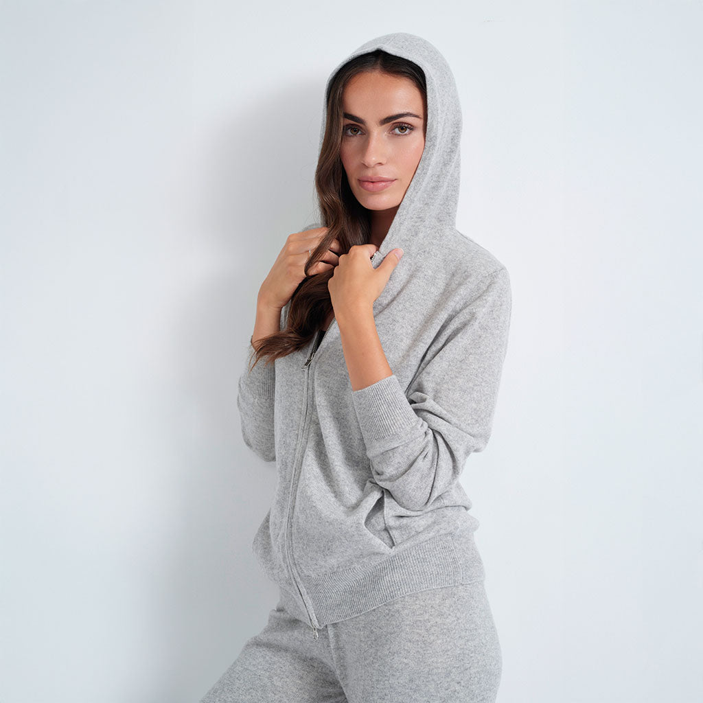 A woman wearing a light gray zip-up hoodie and matching sweatpants, posing against a plain light gray background.