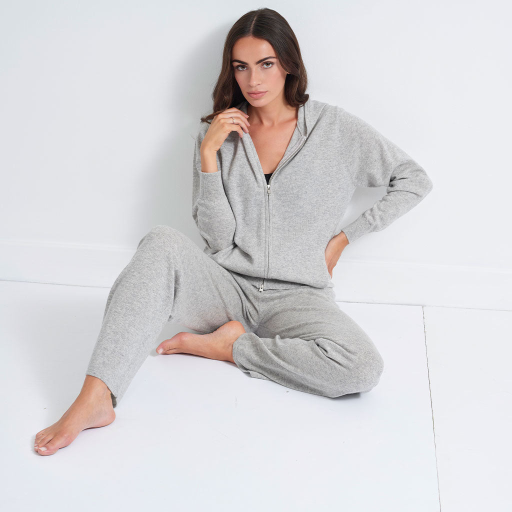 A woman wearing a light gray heathered lounge set consisting of a zip-up hoodie and matching jogger pants, sitting on a white floor against a white wall.