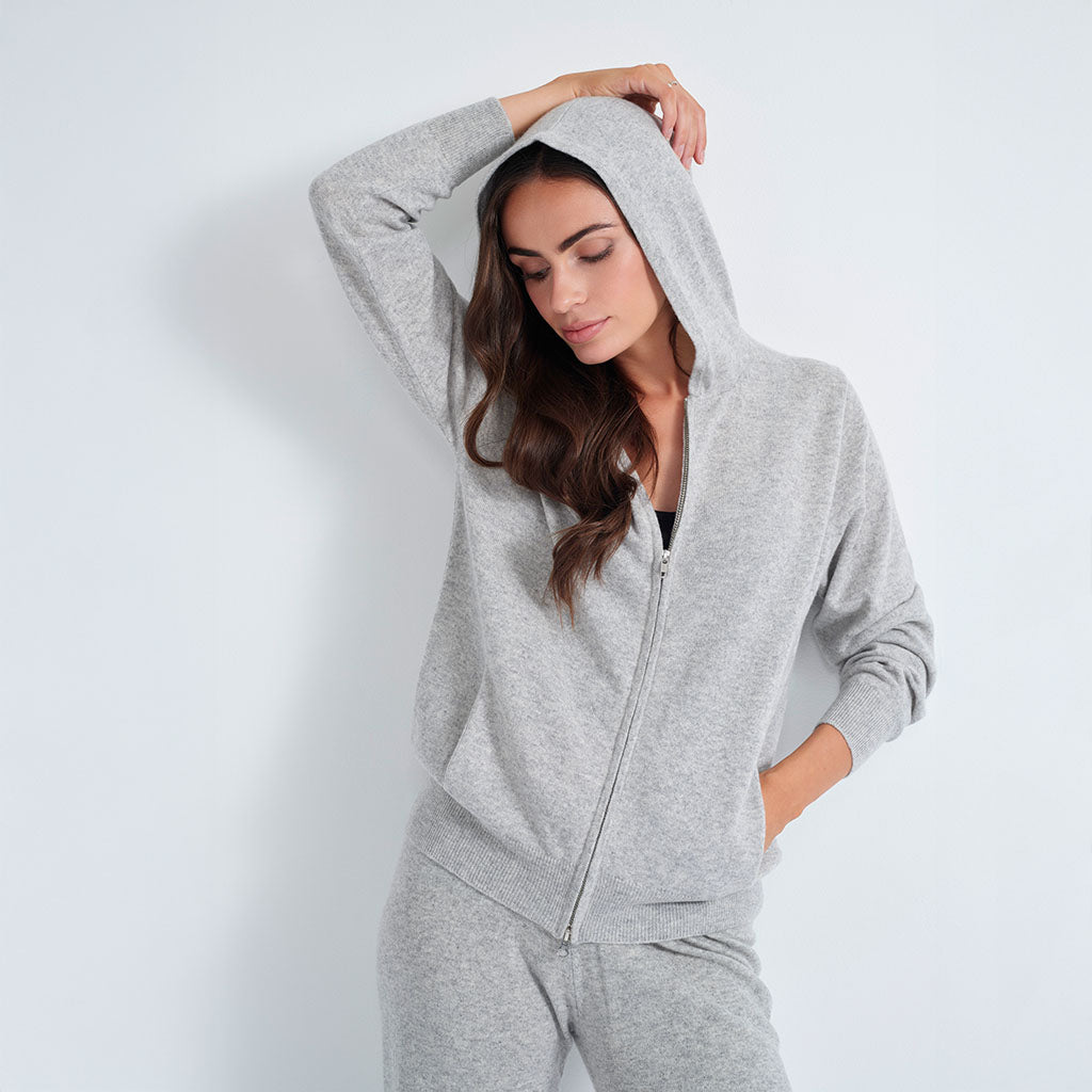 A woman wearing a light gray heather zip-up hoodie and matching sweatpants, posing against a plain light gray wall.