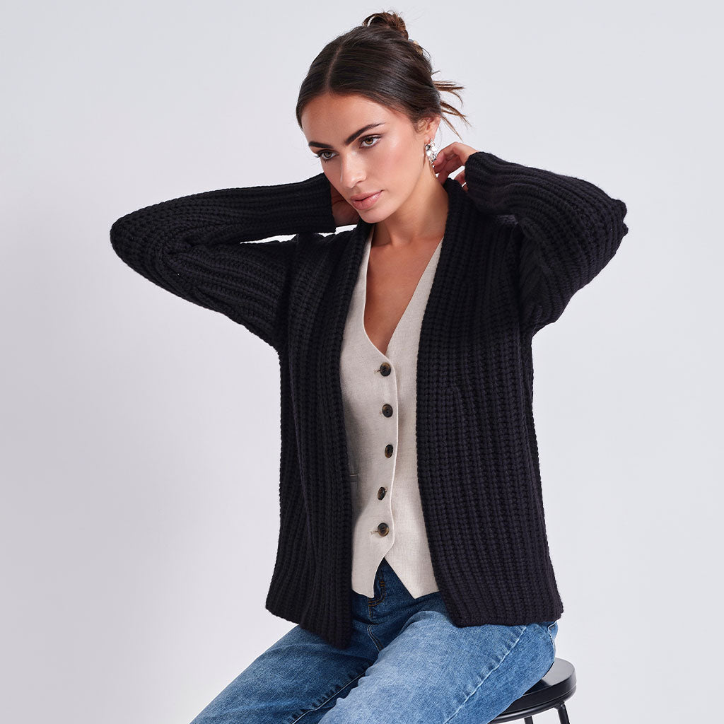 A woman wearing a black ribbed knit cardigan over a cream button-front vest and blue jeans, seated against a light gray studio background.