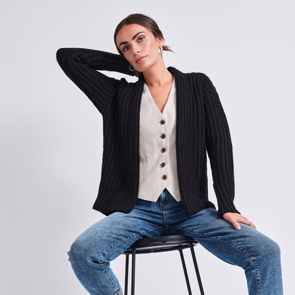 A woman sits on a black stool wearing a black ribbed knit cardigan over a cream button-front vest, paired with blue jeans, against a light gray studio background.