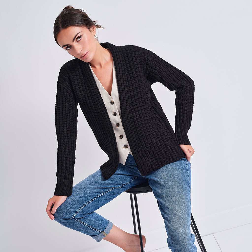 A woman models a black ribbed knit cardigan over a cream button-front vest and blue jeans, seated on a black stool against a light gray studio background.