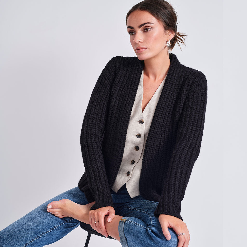A woman seated against a light gray studio background wearing a black textured knit cardigan over a cream button-front top and blue jeans.