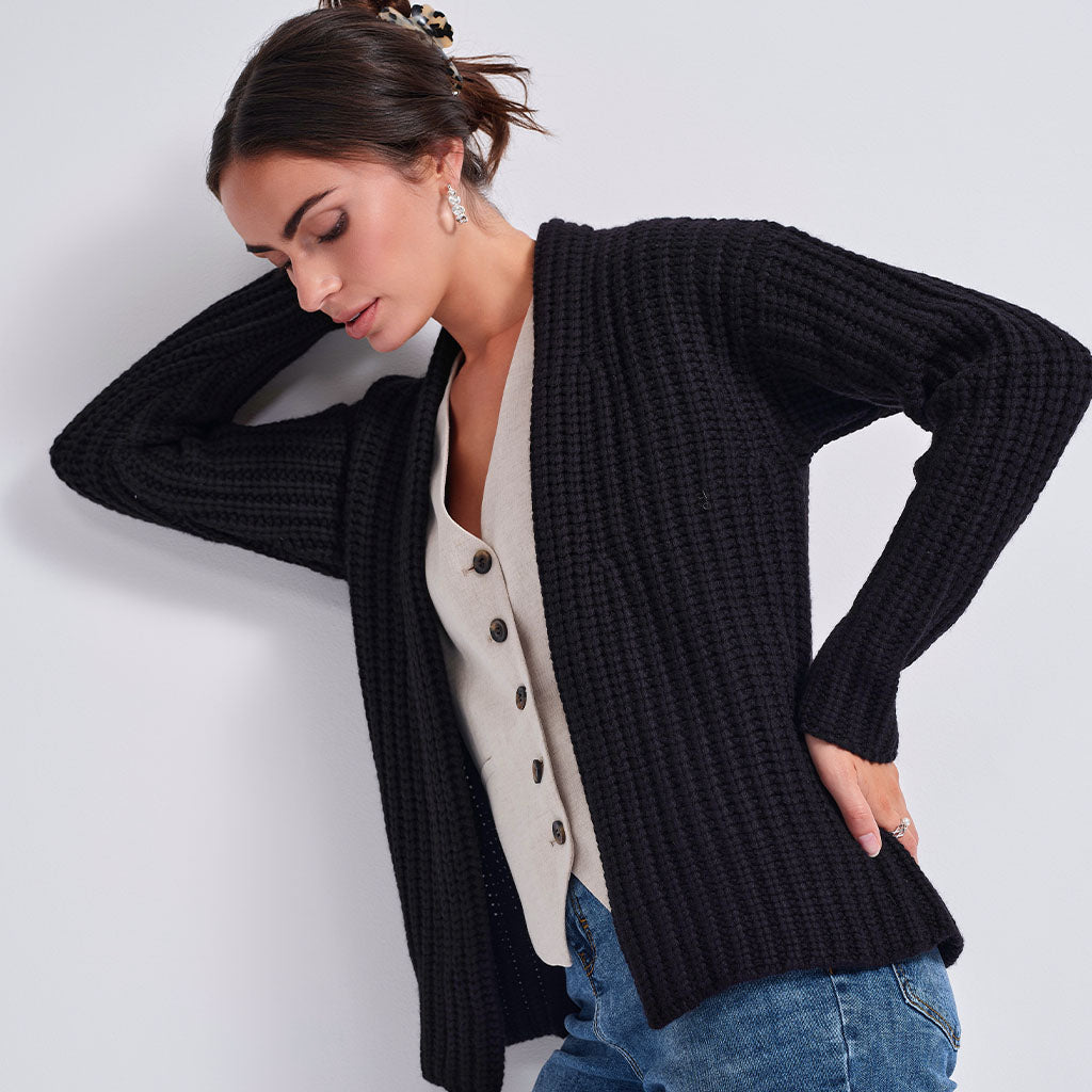 A woman wearing a black ribbed open-front cardigan over a cream button-front top and blue jeans, posed against a light gray studio background.