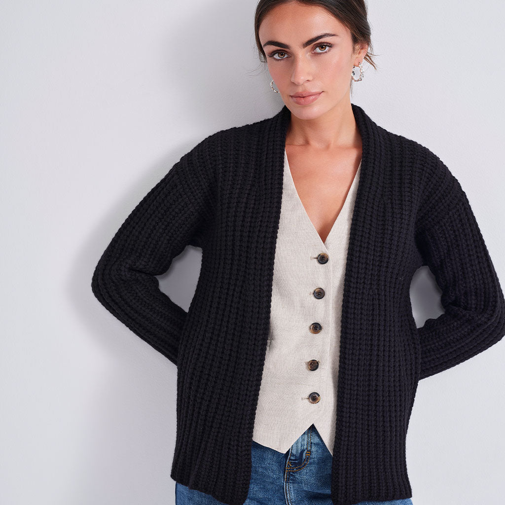 A woman wearing a black ribbed open-front cardigan over a cream button-front vest and blue jeans, standing against a light gray studio background.