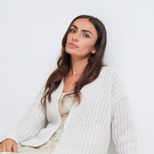 A woman with long brown hair wearing a chunky white waffle-knit cardigan over a light beige patterned dress, photographed against a clean white studio background.
