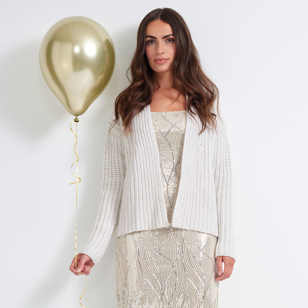 A woman stands against a plain white wall wearing a white textured knit cardigan over a champagne-colored sequined dress, holding a metallic gold balloon.