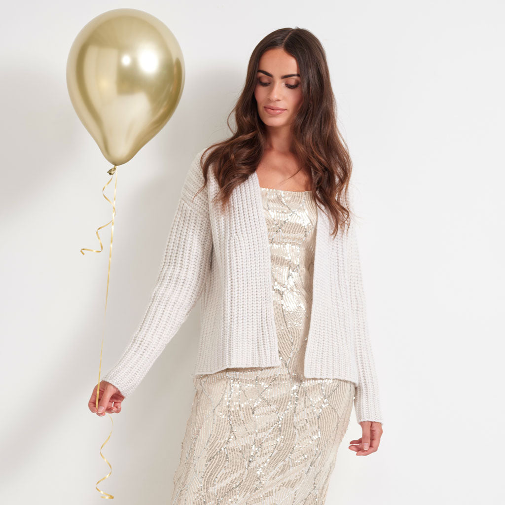 A woman wearing a cream knit cardigan over a champagne sequined dress, holding a metallic gold balloon against a plain white background.