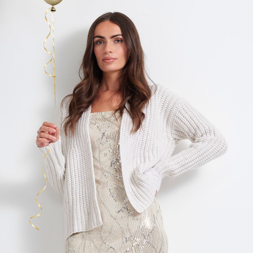 A woman models a white chunky knit cardigan over a champagne sequined dress, holding a gold balloon string against a plain white background.