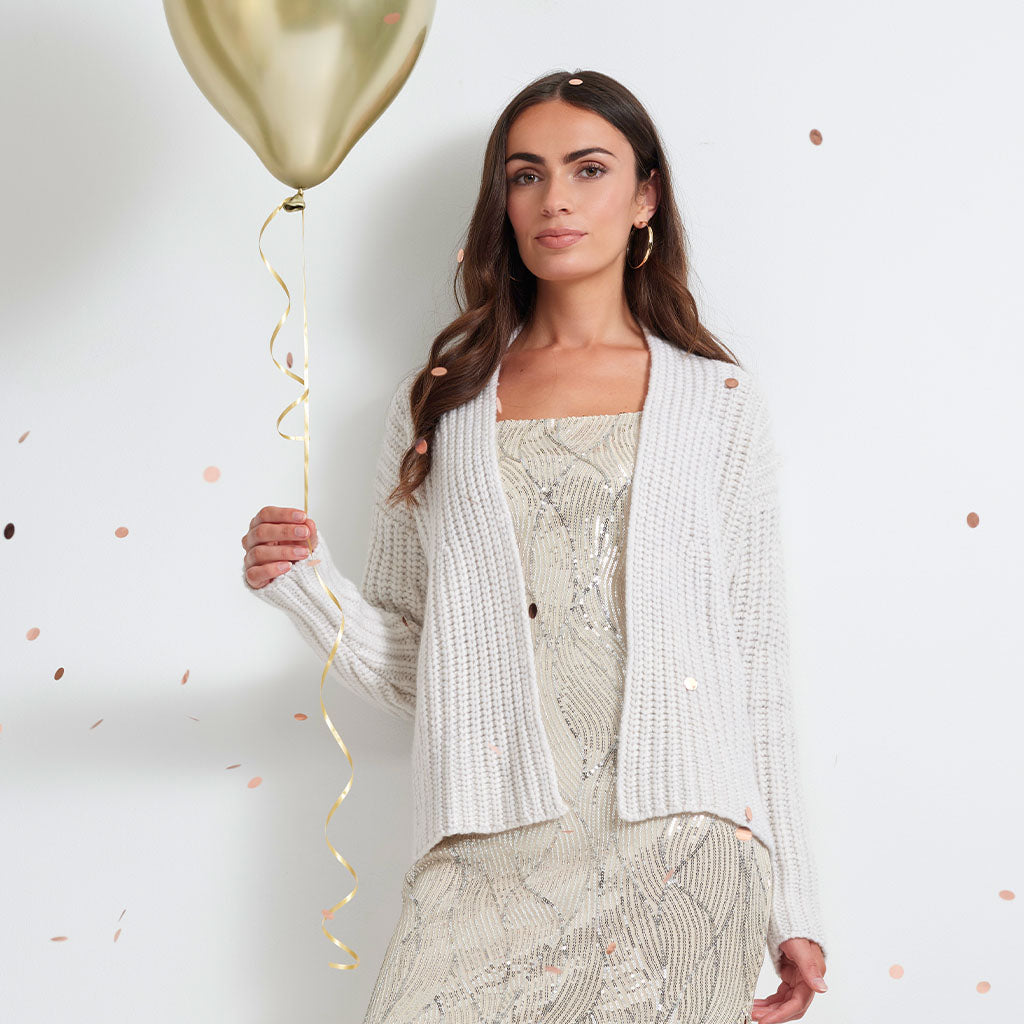 A woman stands against a white wall wearing a white textured knit cardigan over a beige sequined dress, holding a gold balloon with a gold streamer. Rose gold confetti is scattered on the white background.