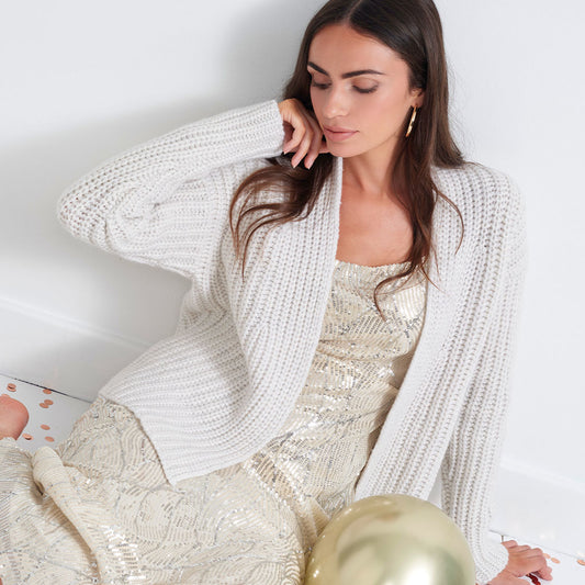 A woman wearing a chunky cream knit cardigan over a champagne sequined dress, sitting against a white wall.