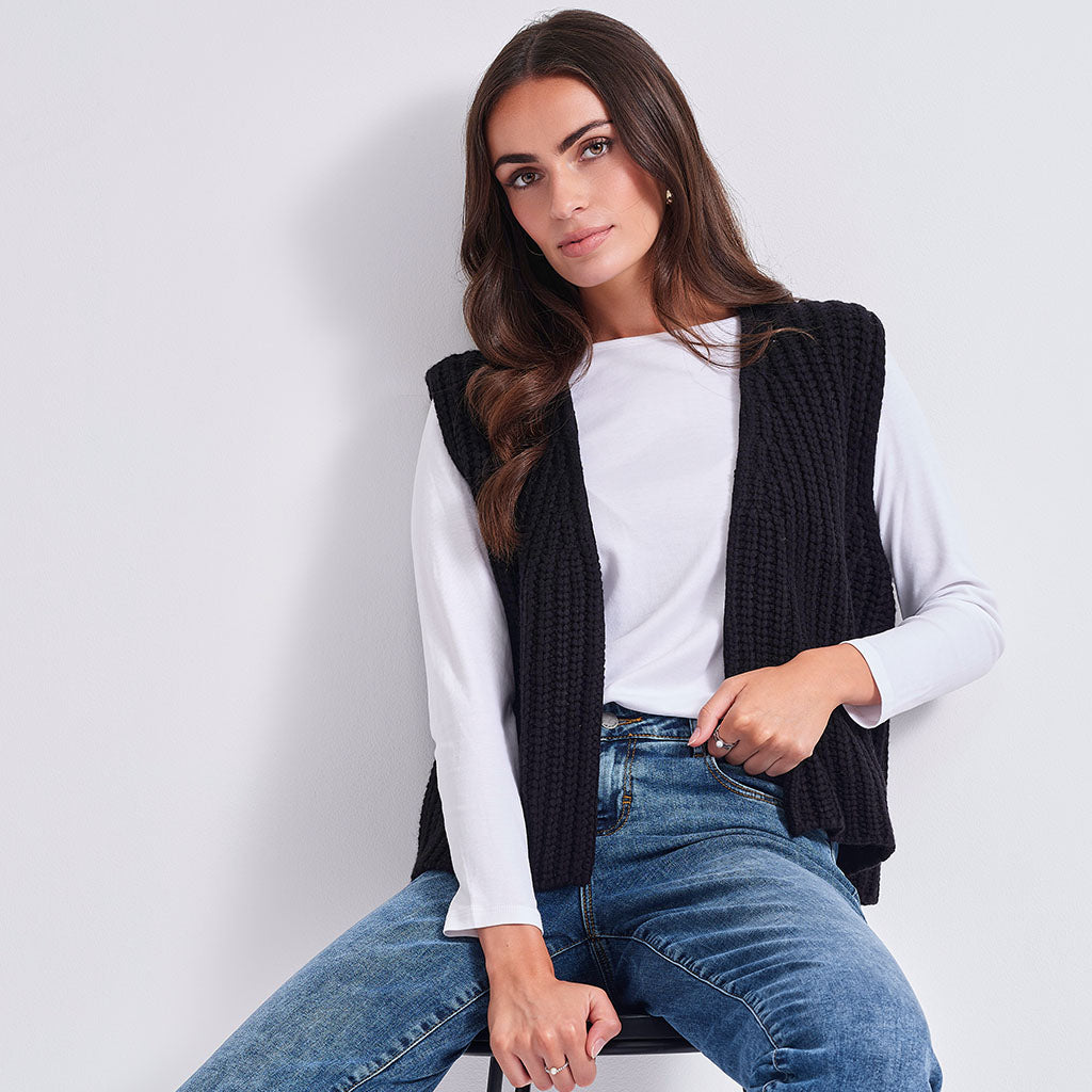 A woman wearing a black ribbed knit sleeveless vest over a white long-sleeve top and blue jeans, seated against a light gray studio background.