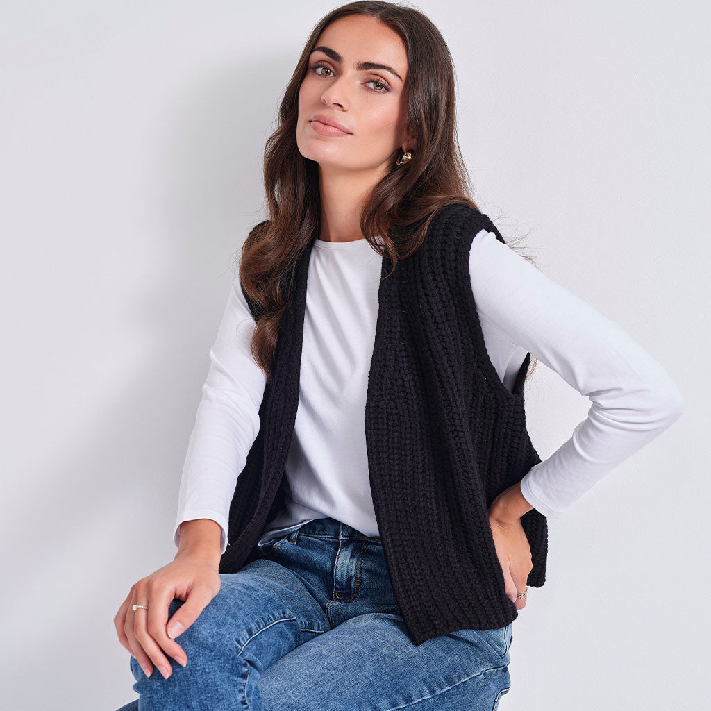 A woman wearing a black ribbed knit sleeveless vest over a white long-sleeve top and blue jeans, seated against a plain light gray background.