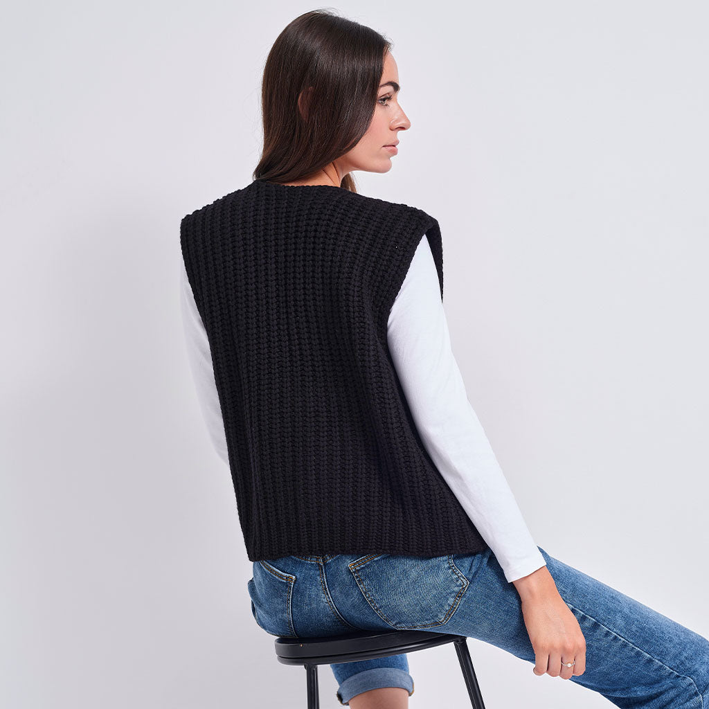 A woman seated on a stool wearing a black ribbed knit sleeveless vest over a white long-sleeve top and blue jeans, photographed against a light gray studio background.
