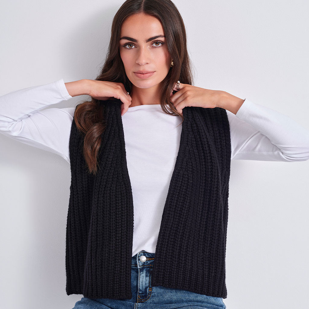 A woman wearing a black ribbed knit sleeveless vest over a white long-sleeve top and blue jeans, posing against a light gray studio background.