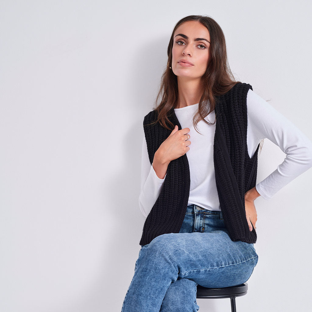 A woman wearing a black ribbed knit sleeveless vest over a white long-sleeve top and blue jeans, seated on a stool against a plain light gray wall.