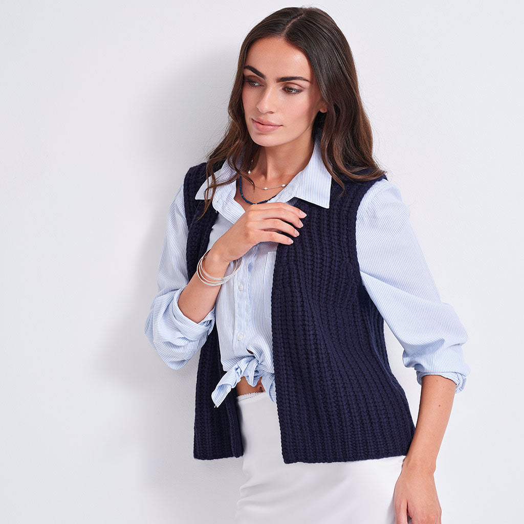 A woman wearing a navy blue ribbed knit vest over a light blue button-down shirt tied at the waist, paired with white pants, against a plain light gray background.