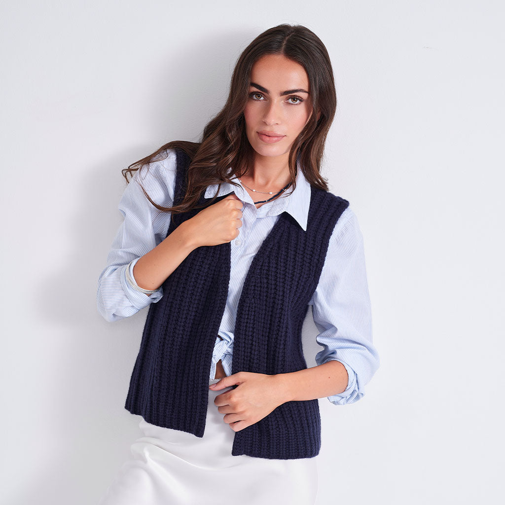 A woman modeling a navy blue ribbed knit sleeveless vest over a light blue button-down shirt and white skirt, set against a plain light gray studio background.