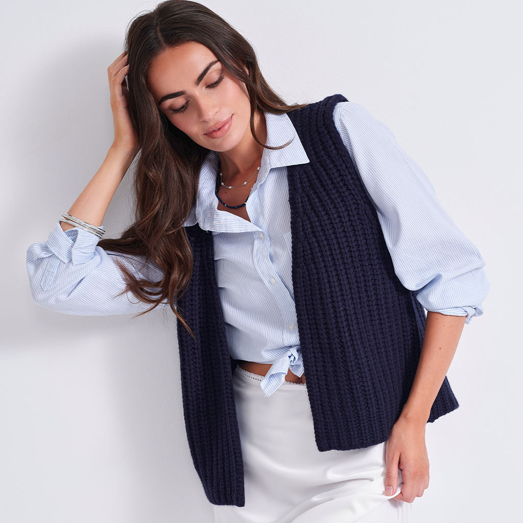 A woman wearing a navy blue ribbed knit sleeveless vest over a light blue and white striped button-down shirt, paired with white pants, set against a plain light gray background.