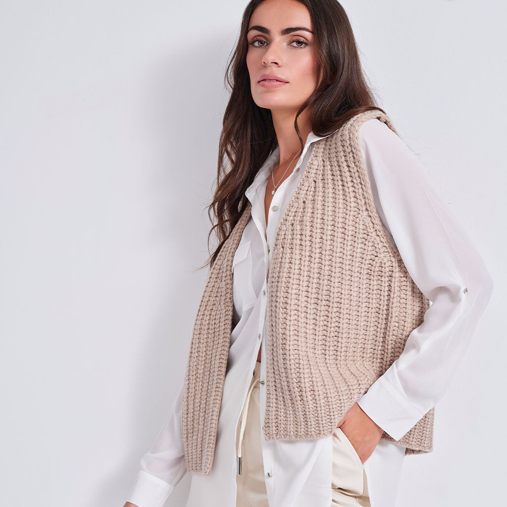 A woman wearing a beige chunky knit sleeveless vest over a white button-up shirt, photographed against a clean light gray studio background.