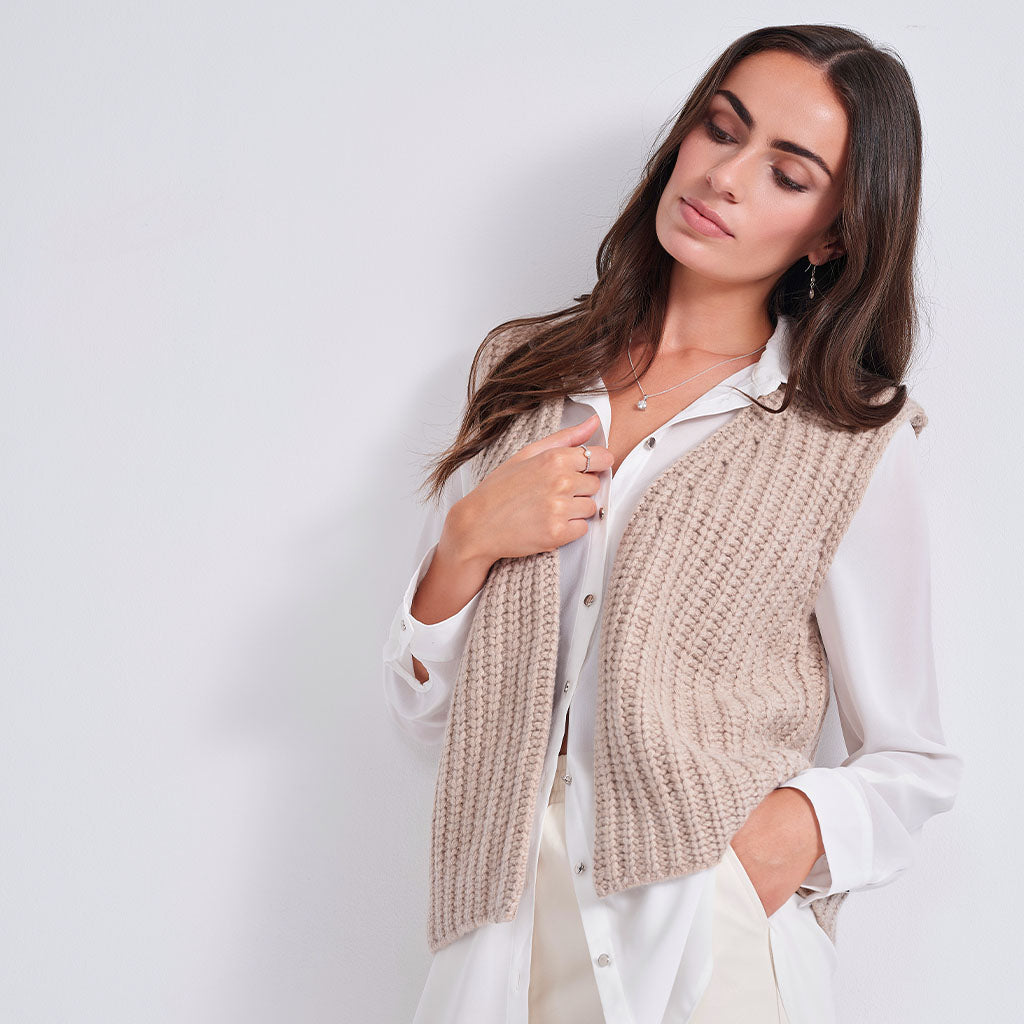 A woman wearing a beige ribbed knit vest over a white button-down shirt, posing against a light gray studio background.