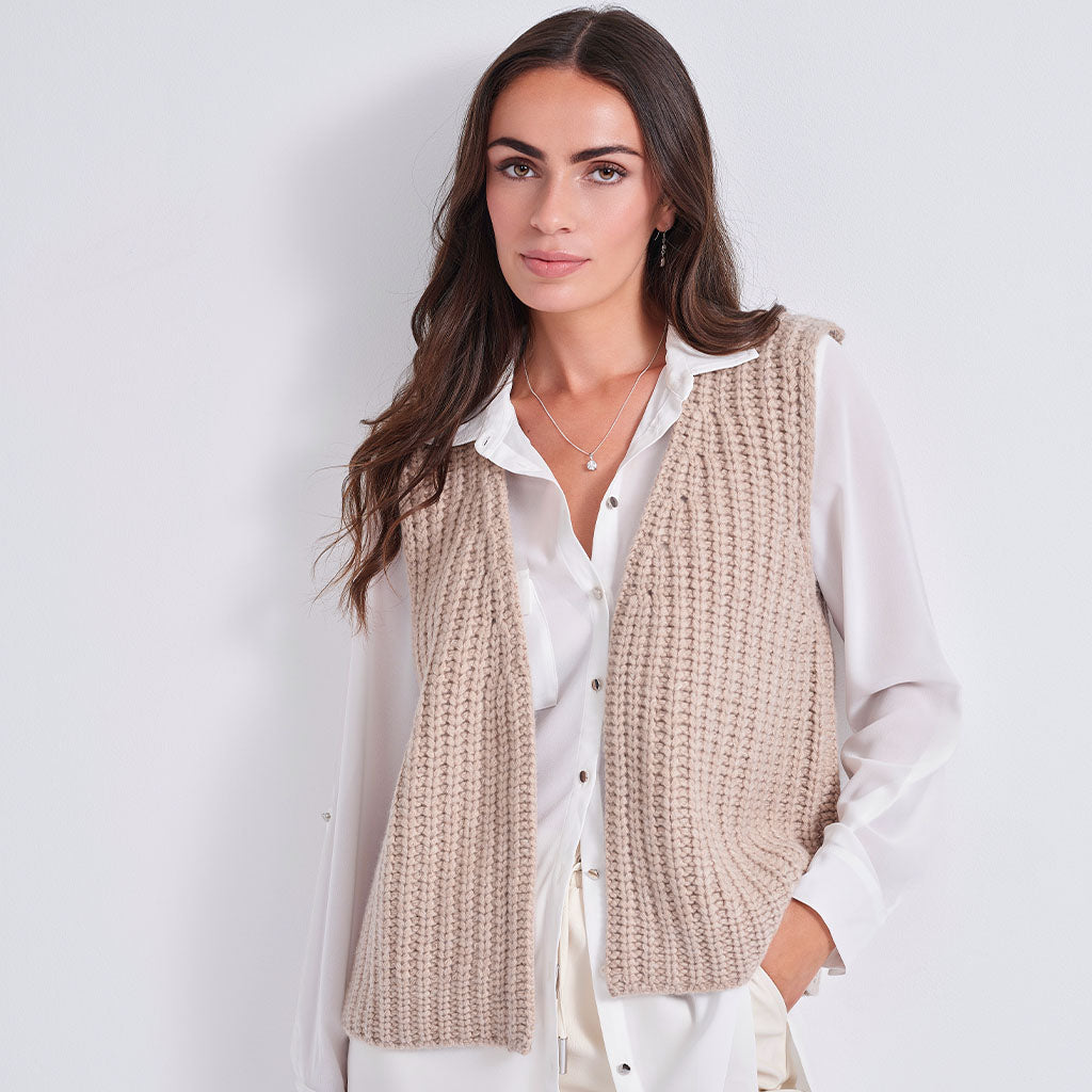 A woman wearing a beige chunky knit sleeveless vest over a white button-down shirt, photographed against a plain light gray studio background.
