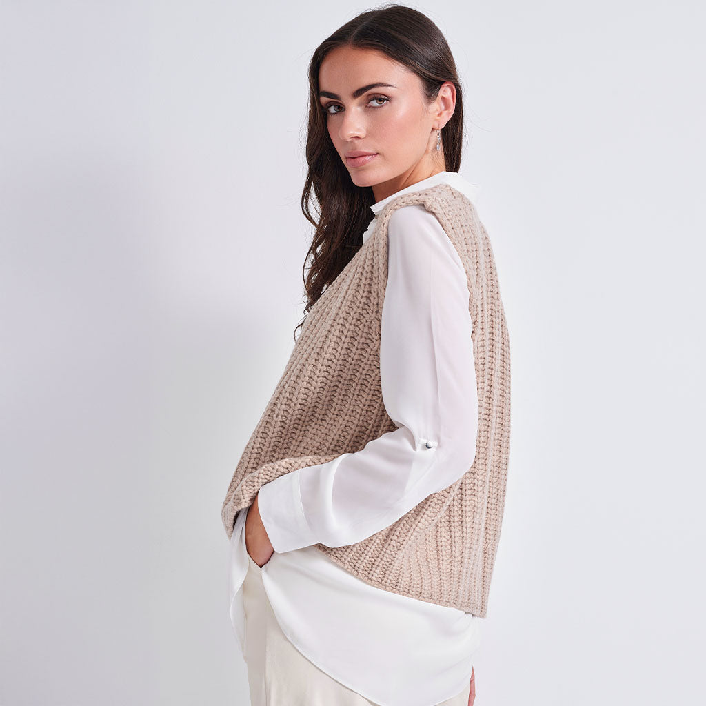 A woman wearing a beige textured knit sleeveless vest over a white long-sleeve blouse, photographed against a clean light gray studio background.